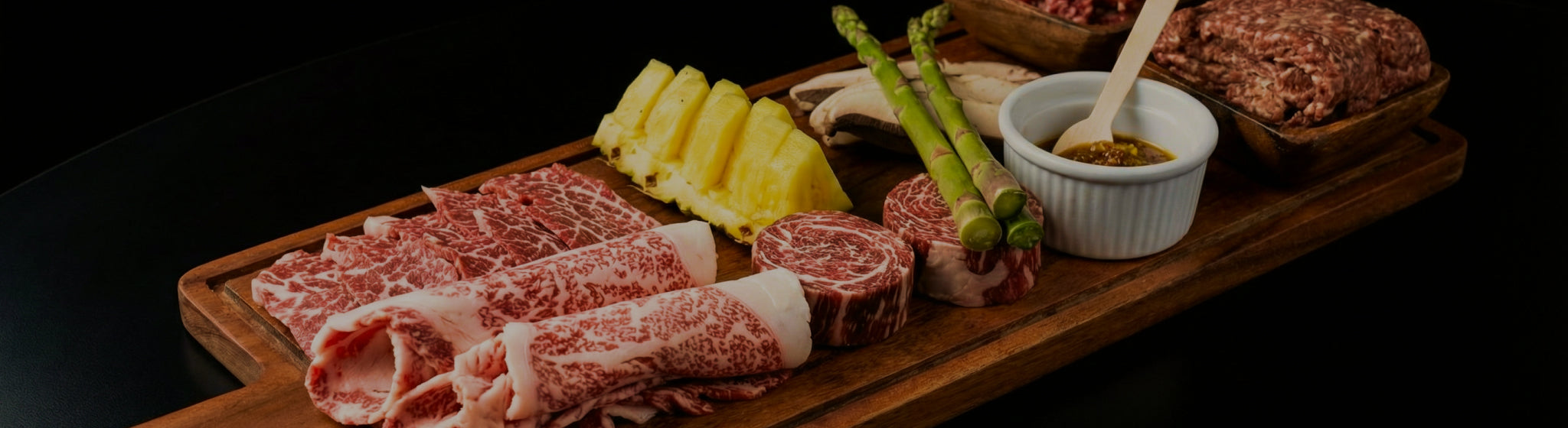 Assorted raw meats on a wooden board with garnishes on a dark background